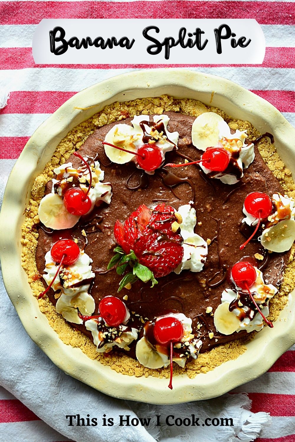 Banana Split Pie Dessert in a round pie dish with nuts, chocolate, strawberries, cherries, and whipped cream on a red and white striped cloth.