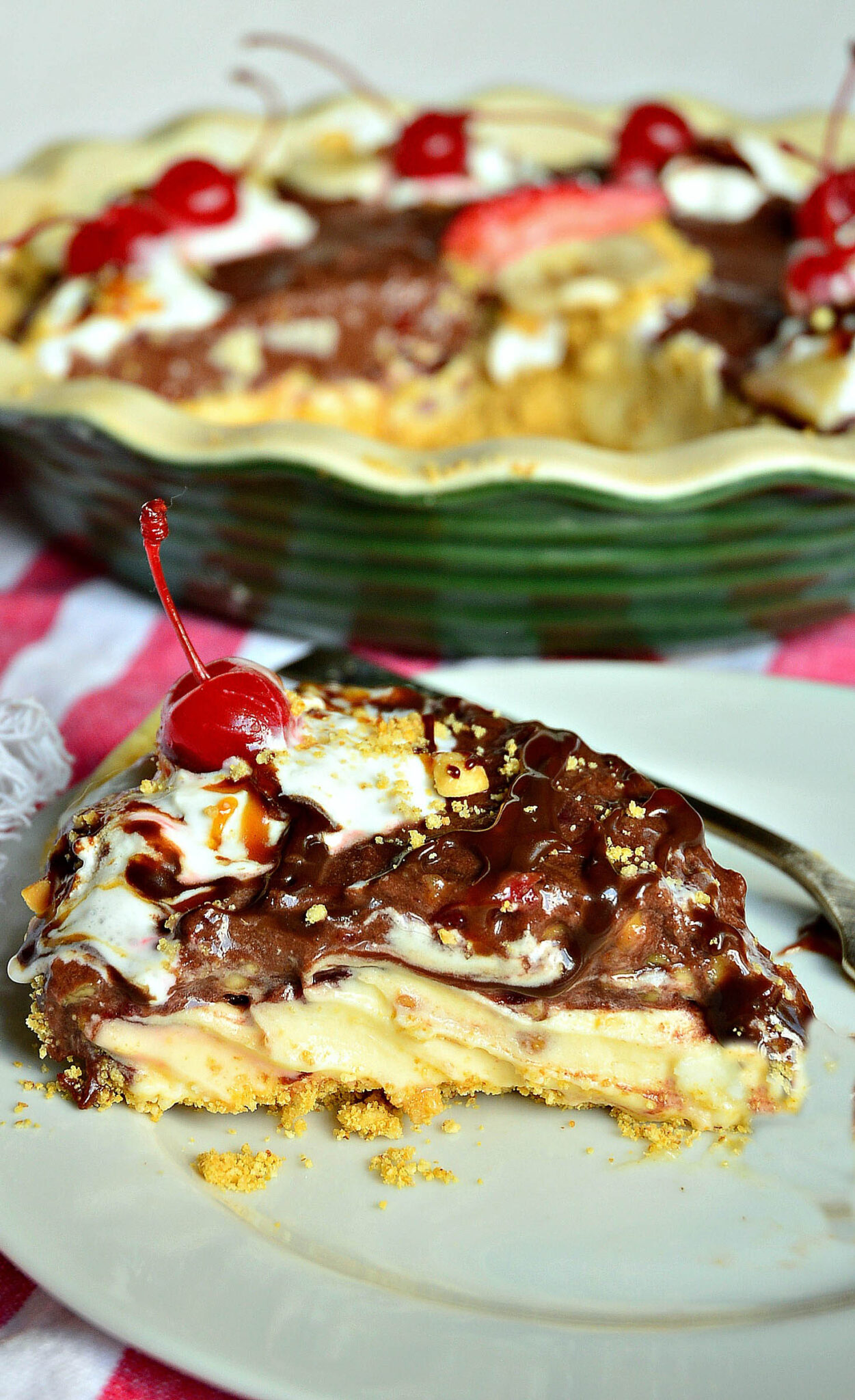 piece of pie with crumble base, creamy middle, and chocolate, nuts, cherry and whipped cream on top with rest of pie in green pie dish in background.