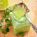 peruvian green sauce in glass jar with silver spoon on wood board with cilantro and lime