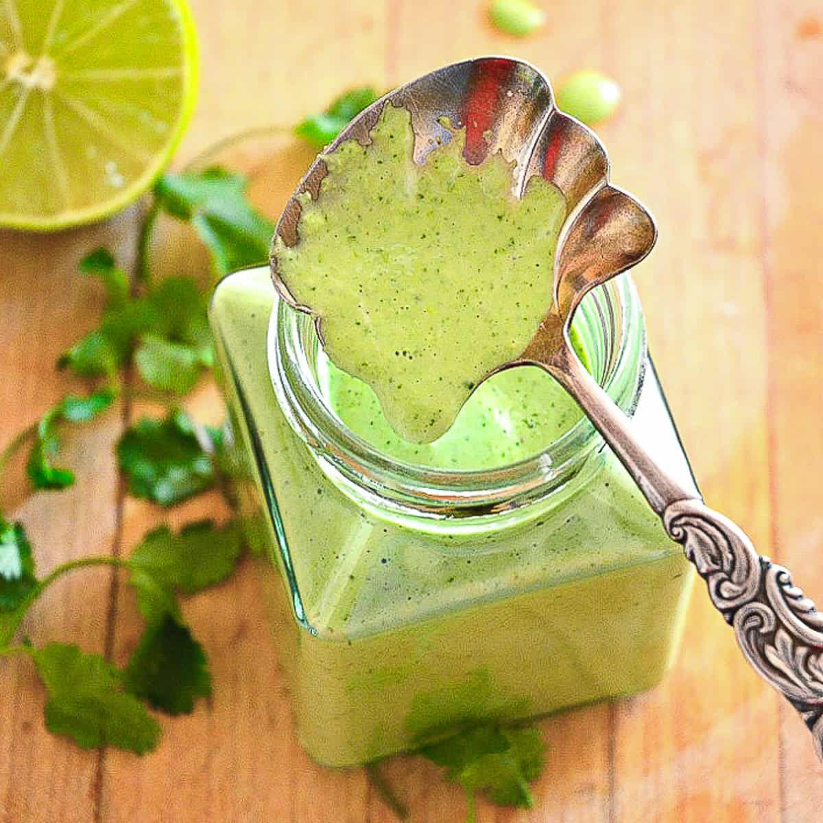 peruvian green sauce in glass jar with silver spoon on wood board with cilantro and lime