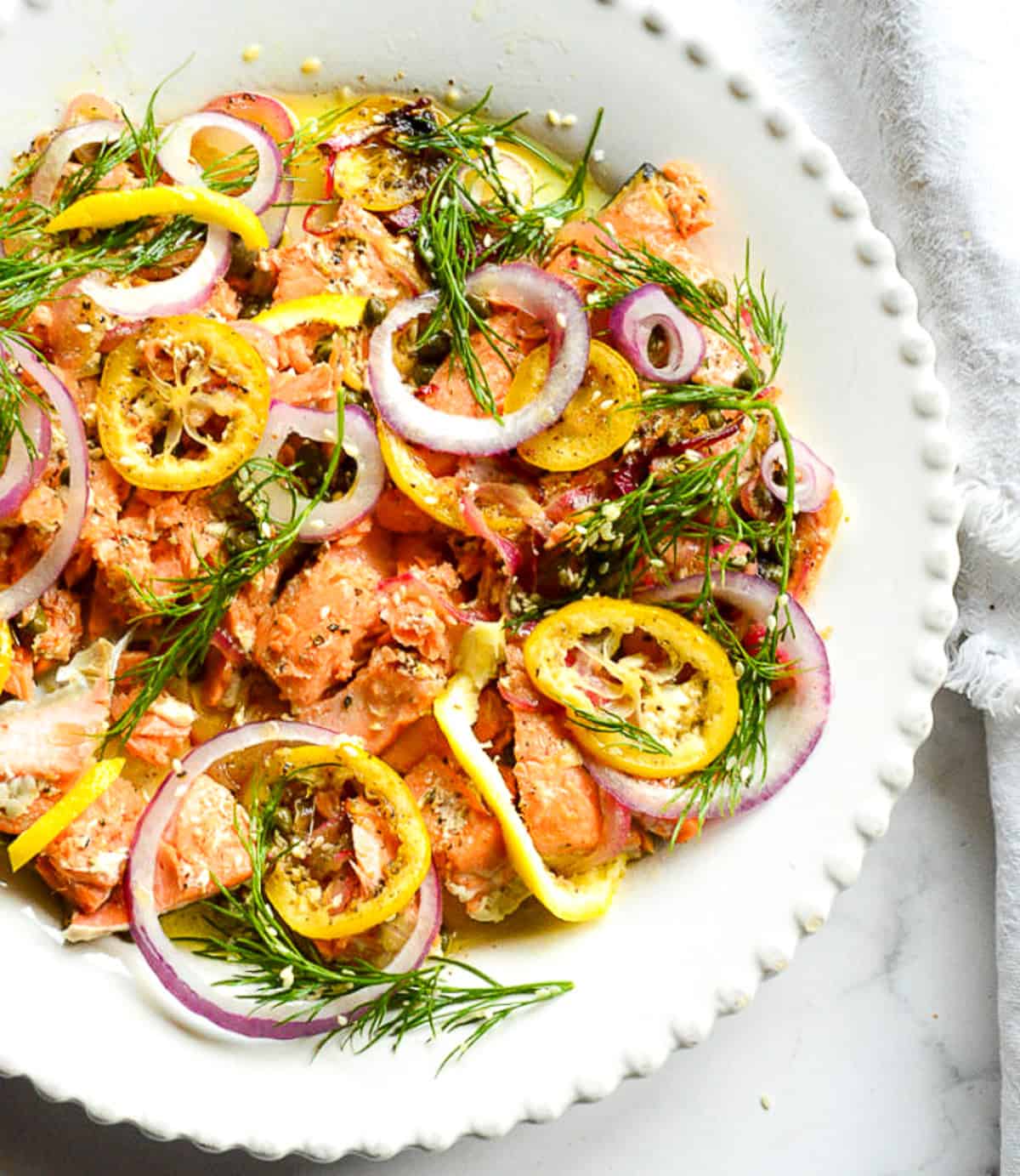 buttered lemon salmon recipe on white plate topped with lemons and red onions and dill