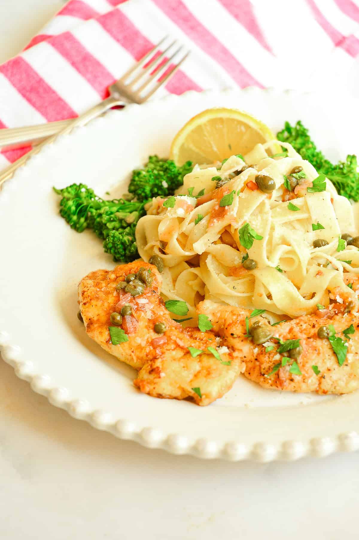 chicken piccata on white plate with noodles and broccolini , capers and lemon