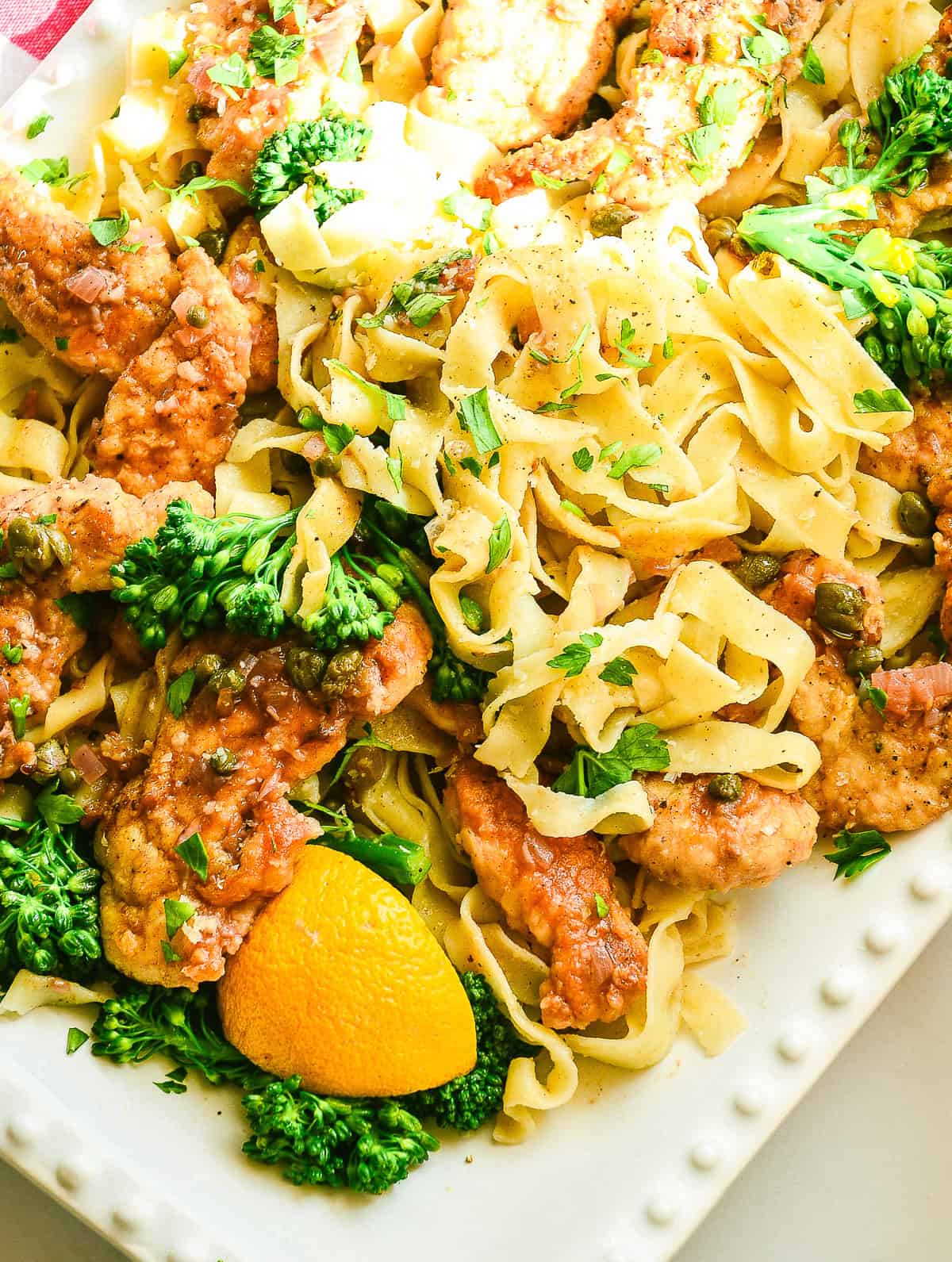 chicken piccata on white platter with noodles and broccolini and capers