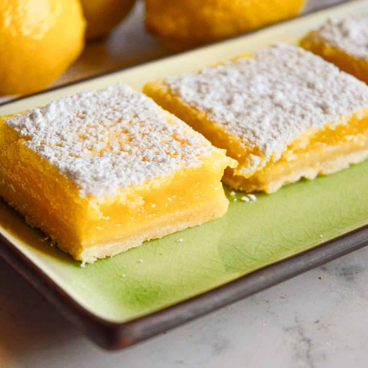 powdered sugar lemon bars on green plate with lemons in background for can you freeze lemon bars