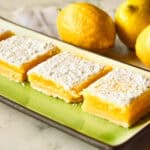lemon bars dusted with powdered sugar on green ceramic tray with lemons in background for can you freeze lemon bars
