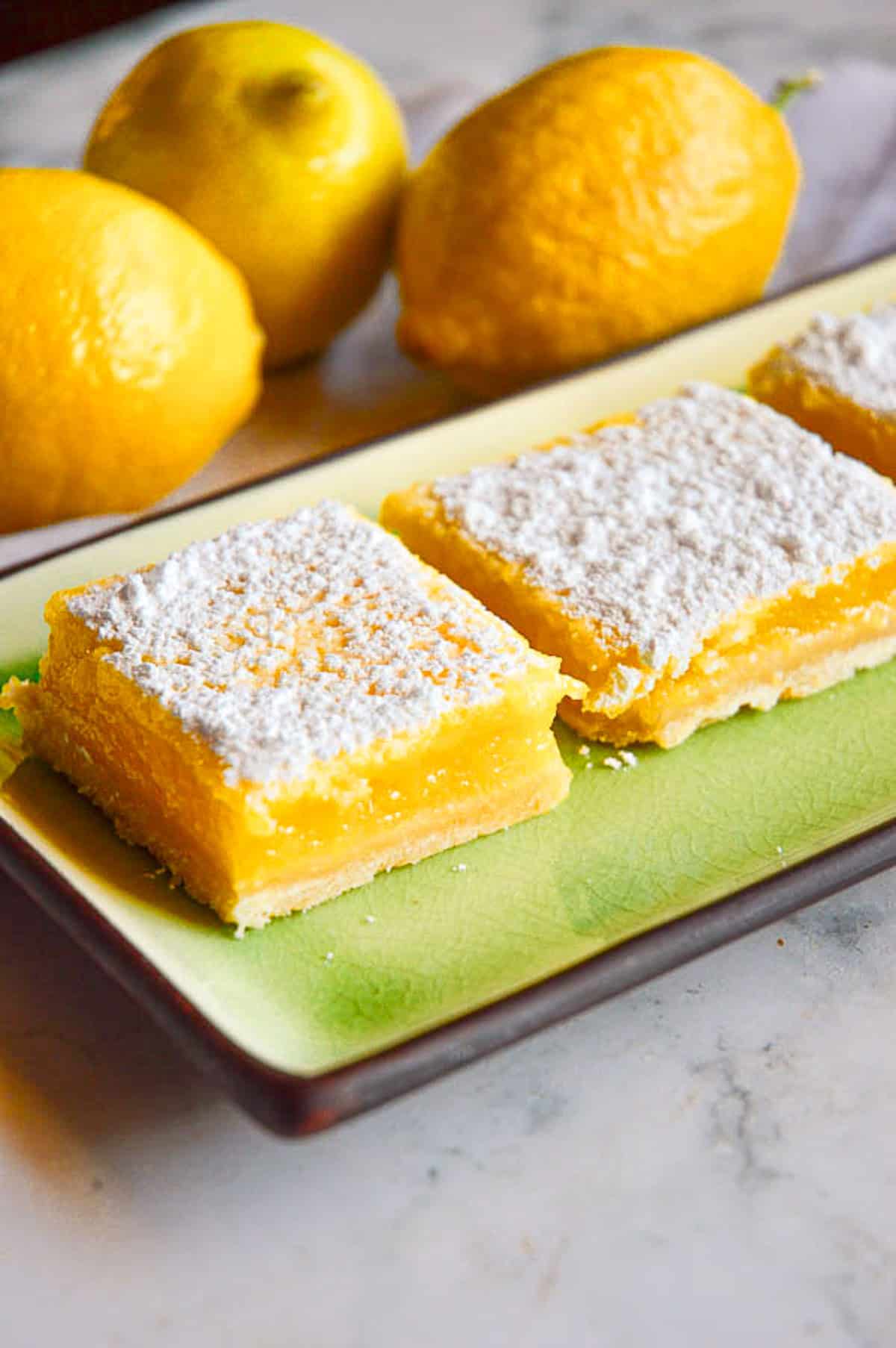 lemon bars on green plate with powdered sugar and lemons in background