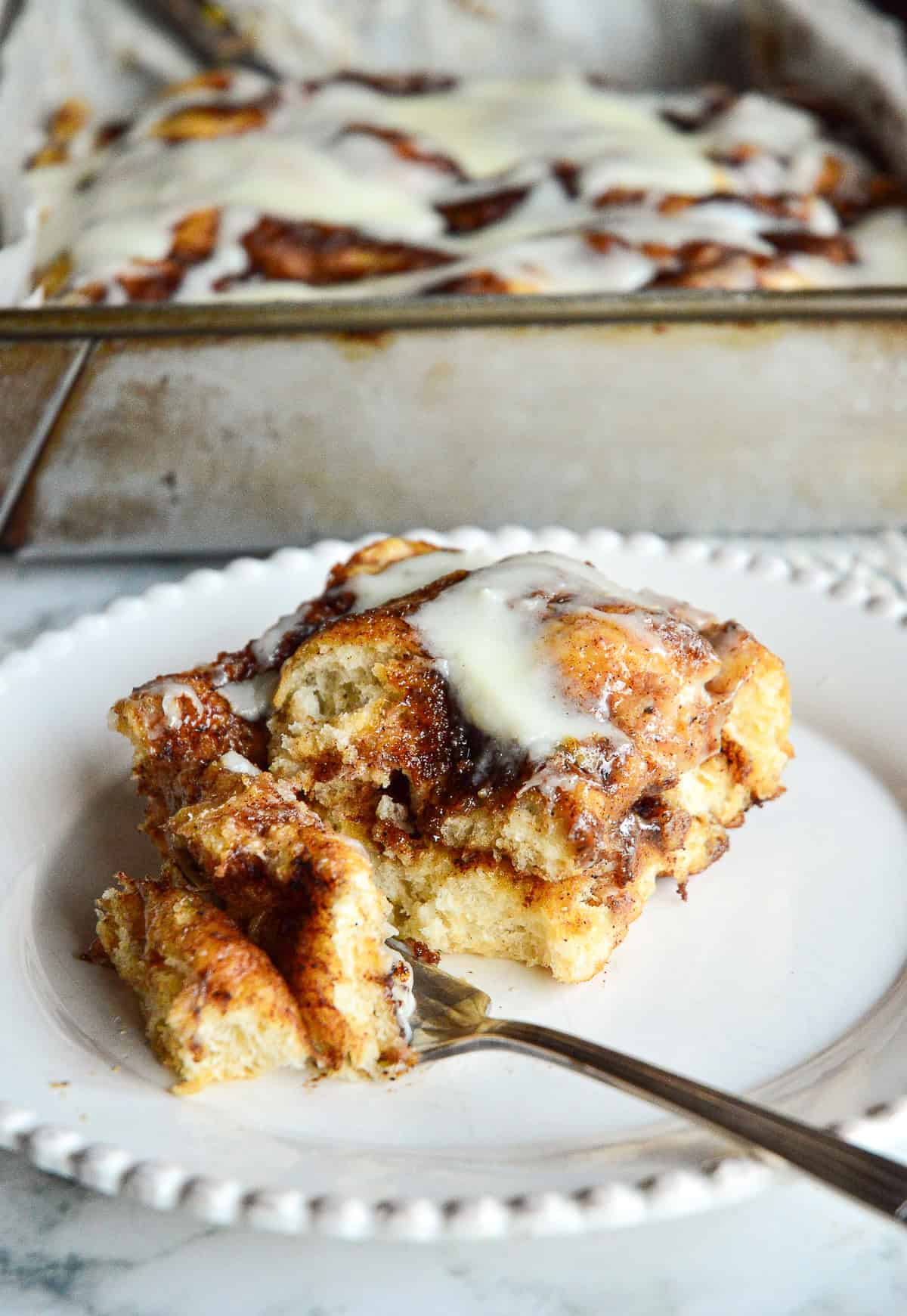 square of cinnamon focaccia bread on white plate with one bite on a fork and a pan of focaccia in the background