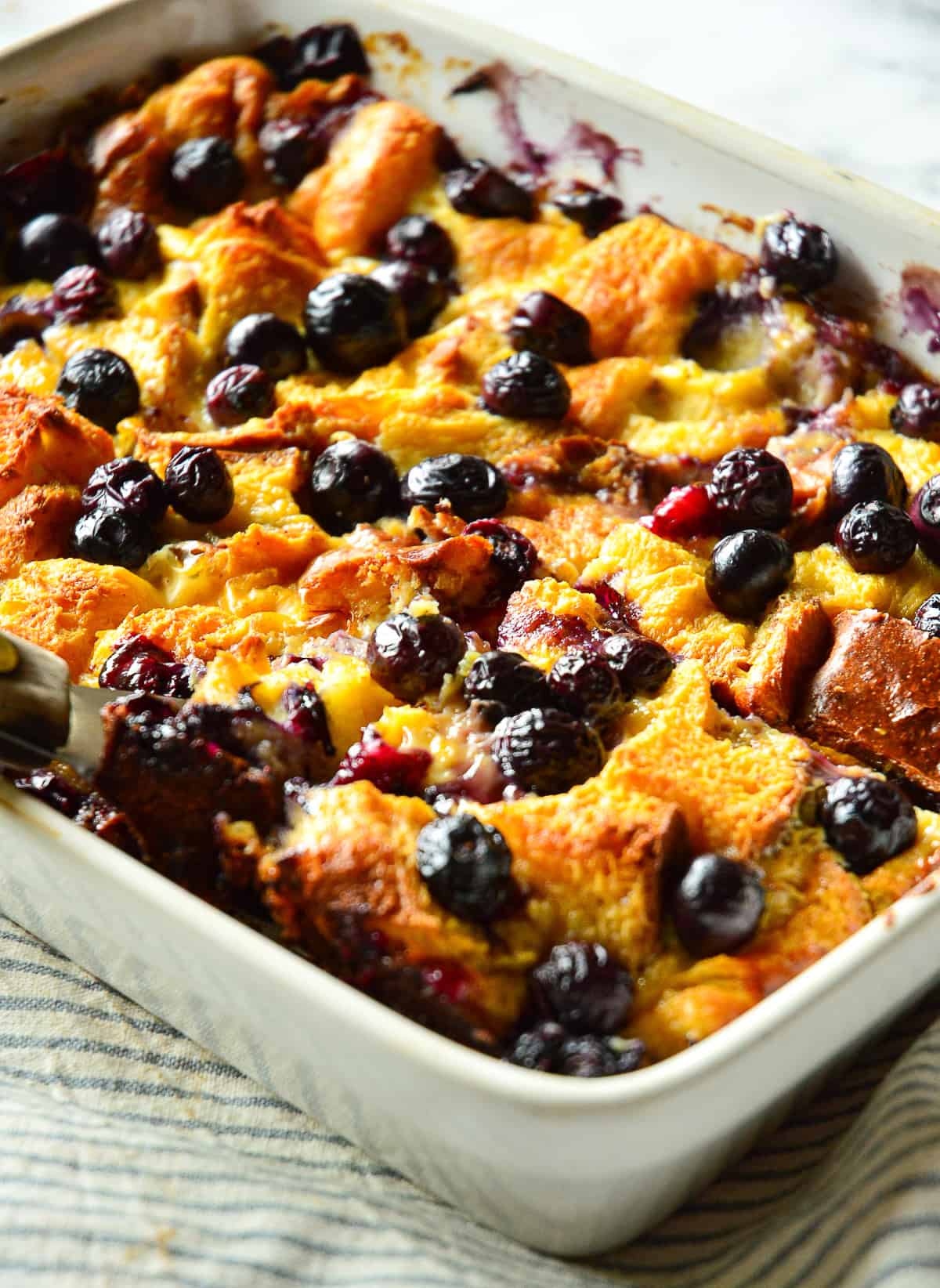 baked french toast casserole with blueberries in white ceramic dish with blue striped napkin