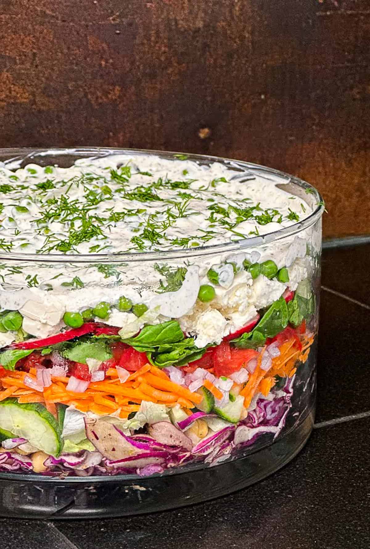 overnight layered salad with red cabbage, chickpeas, cucumbers, shredded carrots, tomatoes, spinach, feta, radishes, peas and za'atar dressing with dill in glass bowl