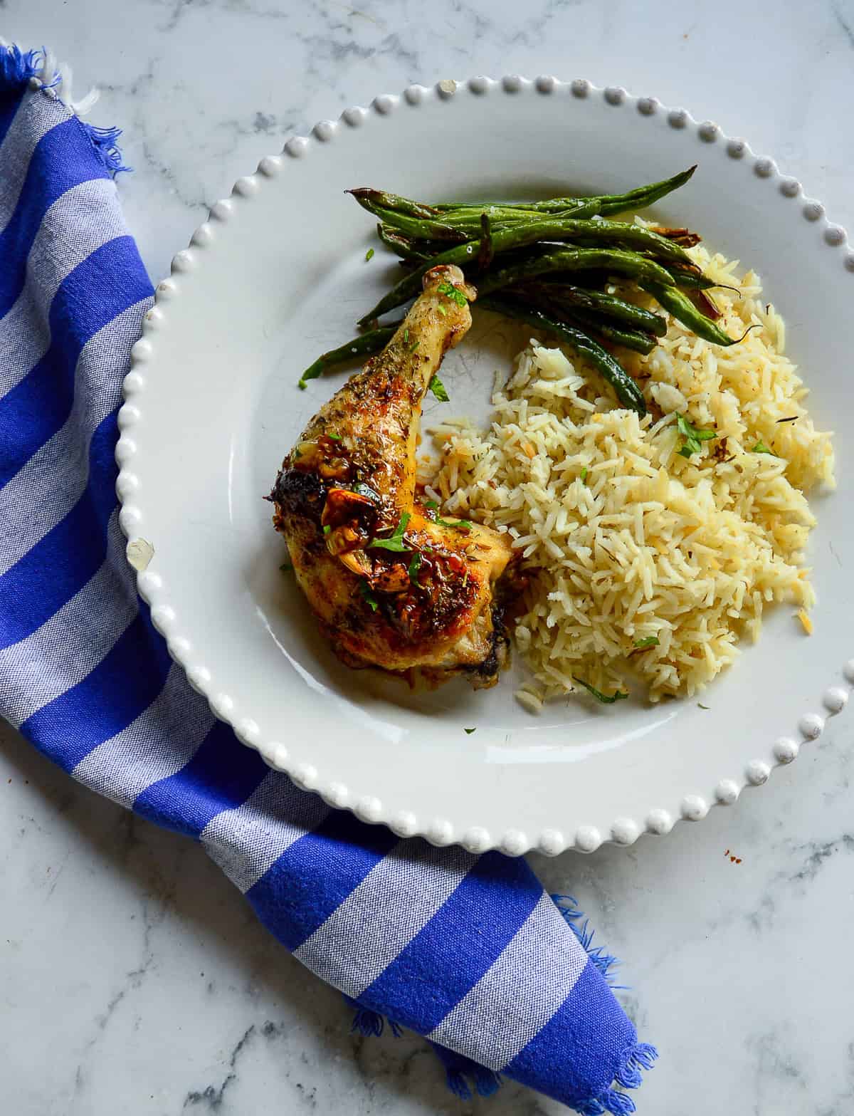dark meat chicken leg on white dinner plate with green beans and rice and a blue napkin on side