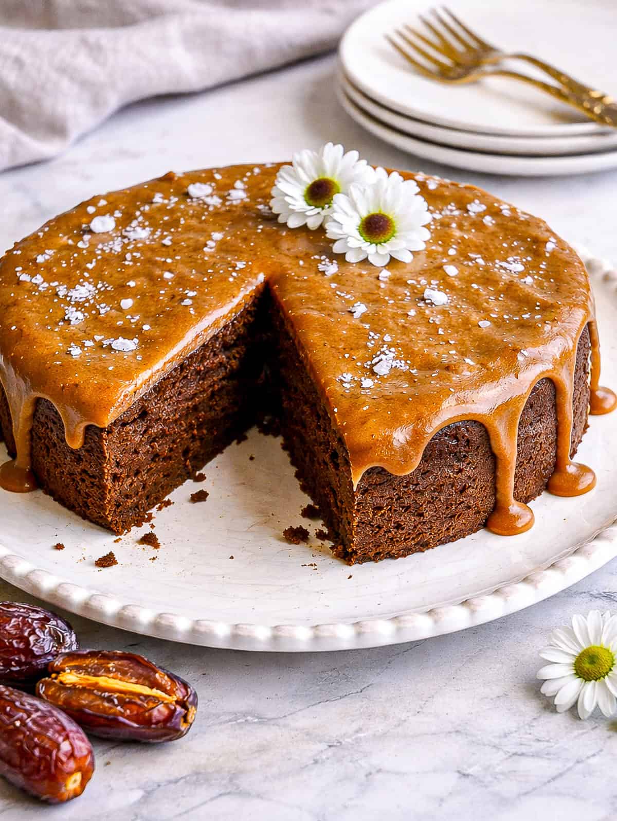 chocolate date cake with caramel frosting on cake plate with flowers and dessert plates and forks in background