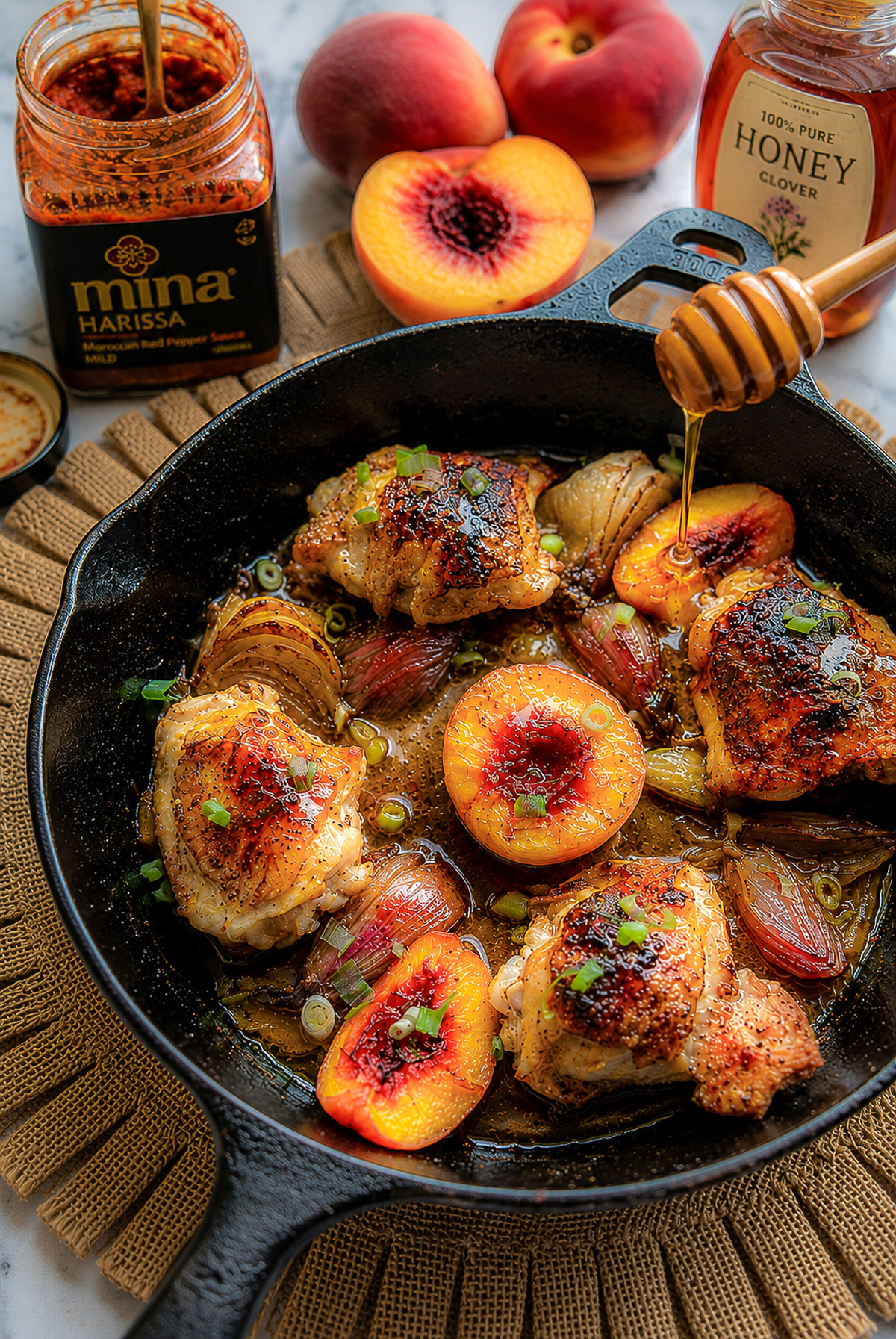 harissa honey chicken in skillet with honey drizzling over the top, fresh peaches and harissa in the background with honey